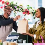 A mid adult African-American man, in his 30s, working at the checkout counter of a gift and flower shop, helping a customer make a purchase. The customer is a young African-American woman in her 20s.